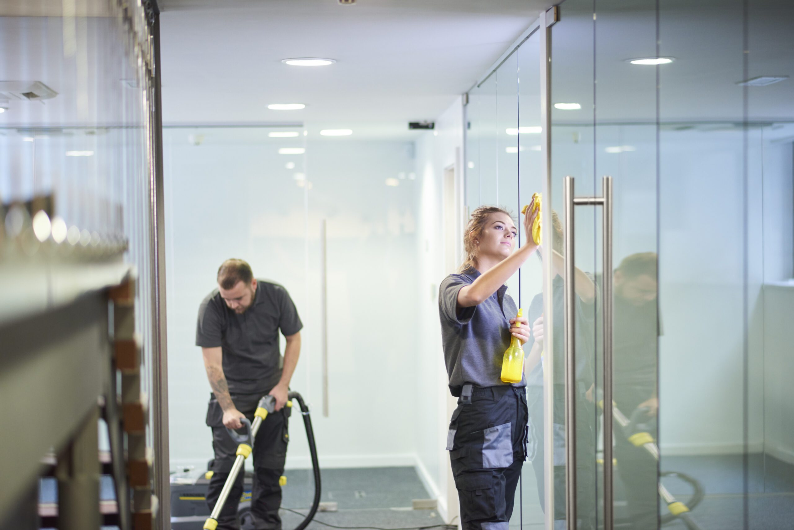 Blog 30 a female cleaning contractor is polishing the glass partition offices whilst In the background a male colleague steam cleans an office carpet in a empty office in between tenants. .